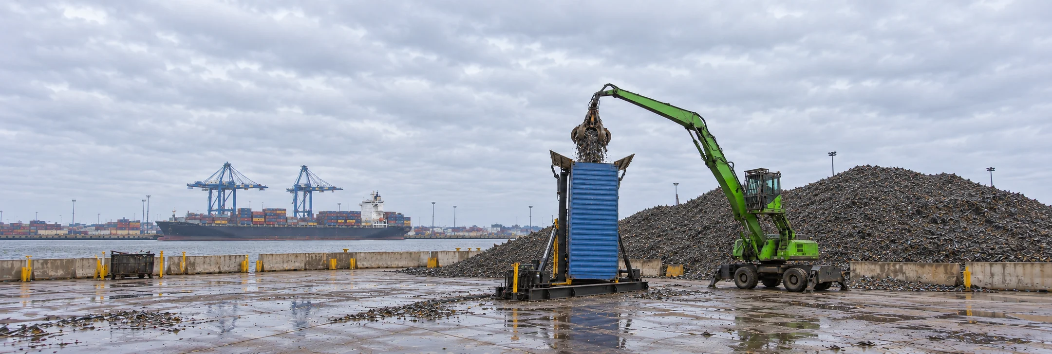 Grab loading shredded scrap into a vertical 20-foot container beside a scrap pile, with a container vessel at the port behind the yard.